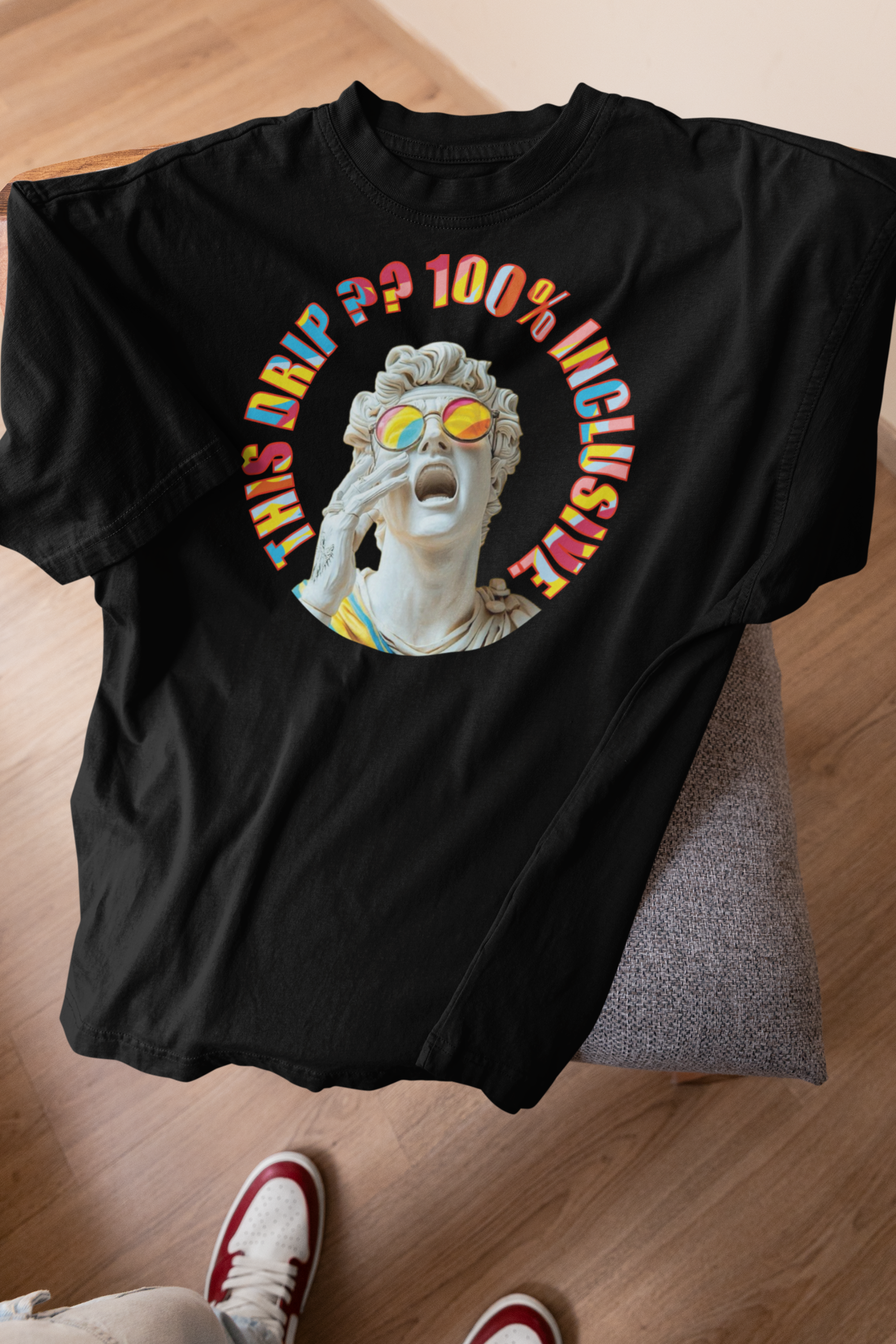 Black ANTI gender-neutral graphic t-shirt laid on a grey seat against a wooden floor, spotlighting a dramatic white marble statue with rainbow-tinted glasses and rainbow-hued lettering that reads “THIS DRIP?? 100% INCLUSIVE.” A bold, pride-centric fashion statement designed for queer-forward and expressive streetwear lovers.

