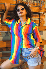 A gender-fluid individual with curly hair and small sunglasses strikes a confident pose, leaning against a wooden slatted wall. They are wearing ANTI's eye-catching short-sleeved shirt, prominently featuring vertical rainbow stripes, styled with denim shorts and a distinctive gold chain belt, showcasing authentic queer fashion.