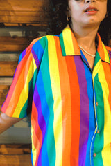 Close-up detail of ANTI's short-sleeved shirt, highlighting its rich, vertical rainbow stripe pattern in vibrant hues of red, orange, yellow, green, blue, and purple. The fabric texture is visible, along with a pearl necklace, emphasizing the shirt's quality and bold, inclusive design