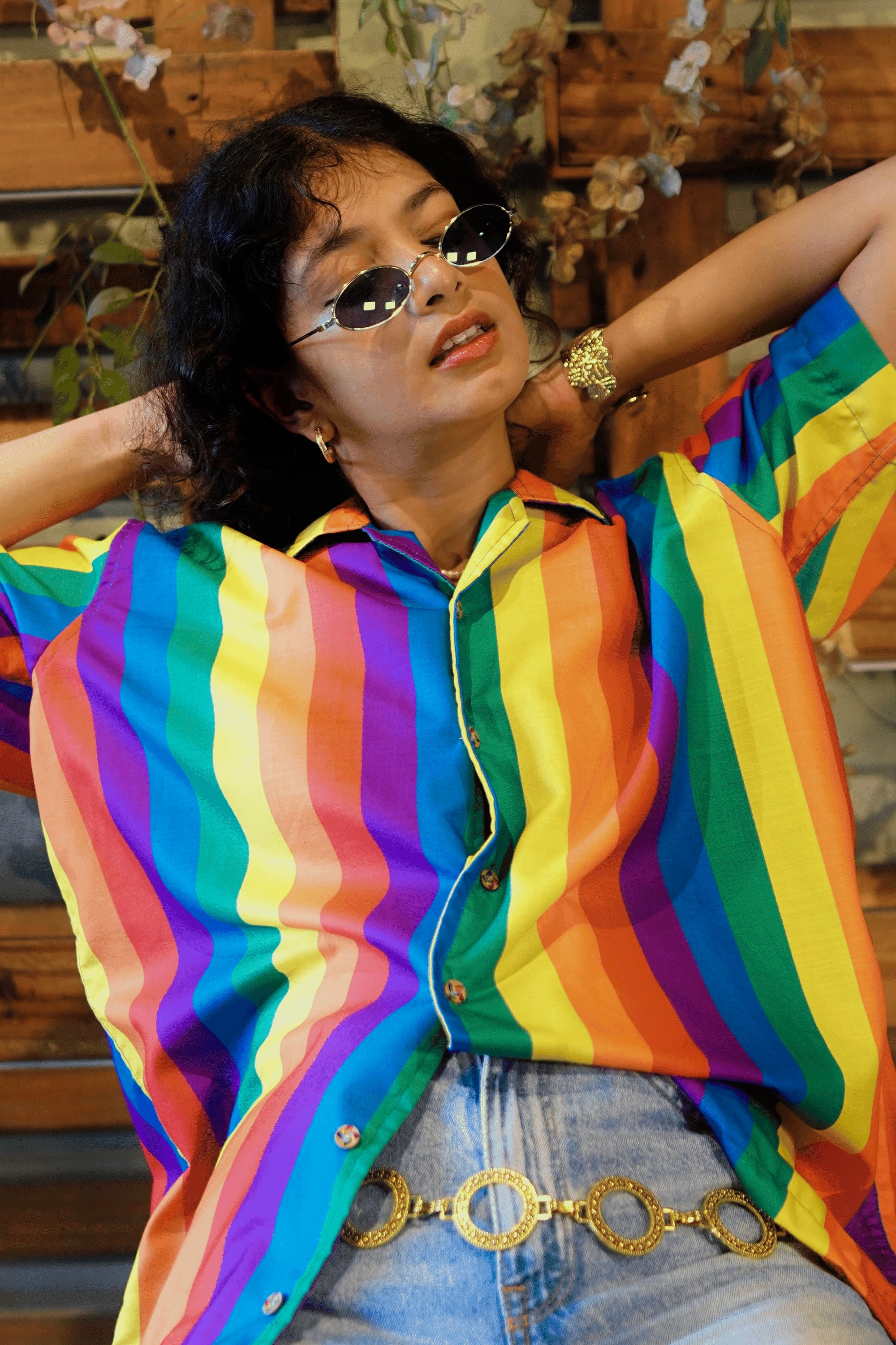 A playful gender-fluid person with curly hair, round sunglasses, and a pearl necklace smiles widely with their tongue out, hand behind their head. They are proudly wearing ANTI's short-sleeved shirt adorned with bold vertical rainbow stripes, perfectly capturing the brand's vibrant, inclusive, and expressive identity.