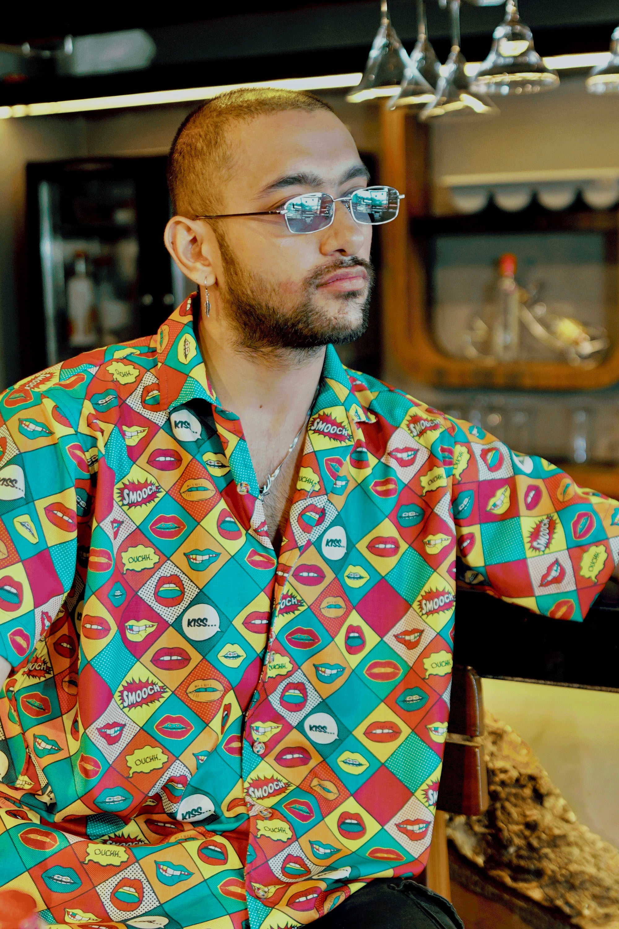 A contemplative gender-fluid individual with cool, tinted glasses and a beard looks off to the side, wearing ANTI's short-sleeved shirt with a striking pop art-inspired design. The shirt is covered in a grid of multi-colored lips and comic book sound effects like 'KISS...' and 'OUCH!,' perfect for a bold, inclusive look.