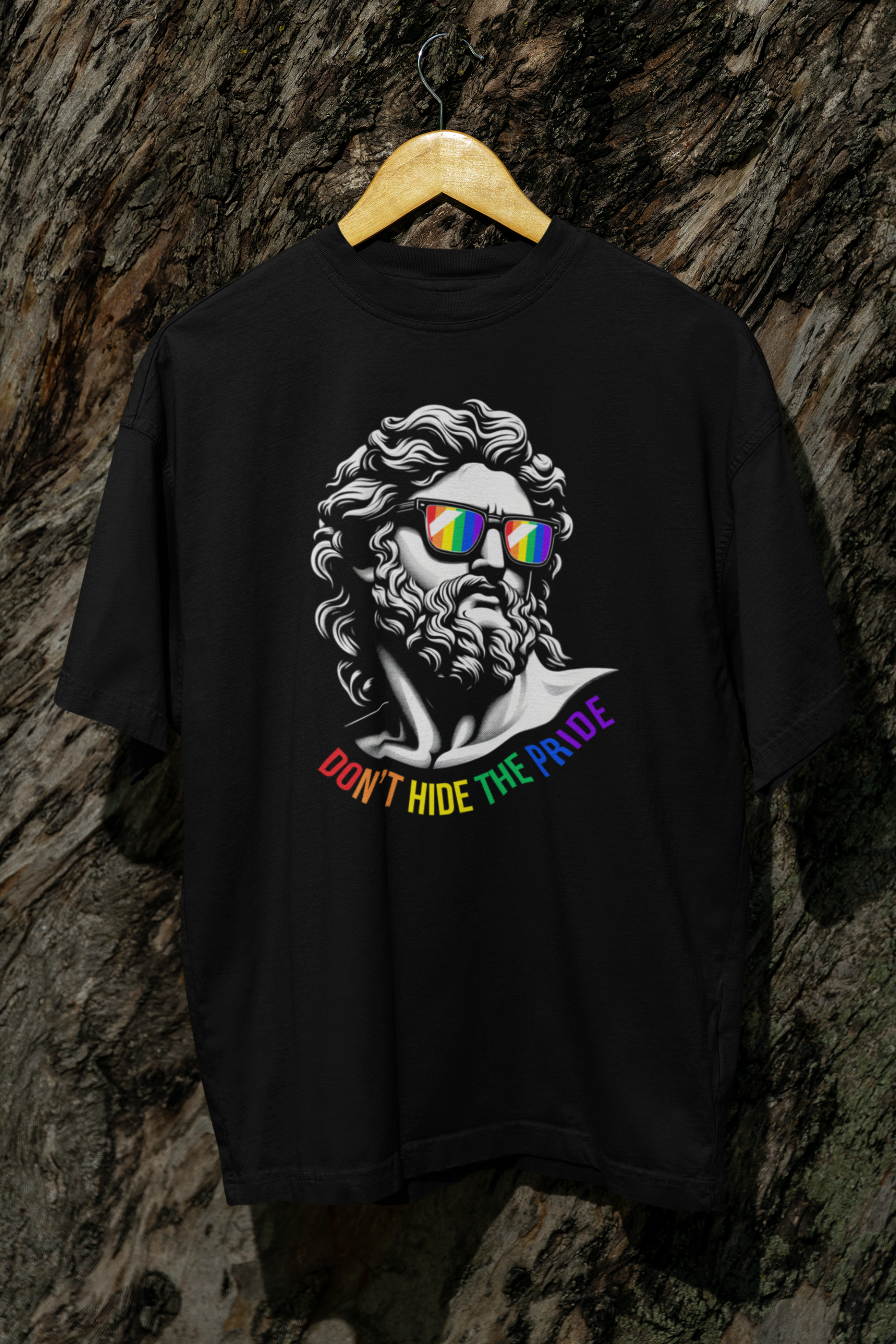 “ANTI’s black ‘Don’t Hide the Pride’ T-shirt hanging on a wooden hanger against a tree, showcasing a queer-positive design of a Greco-Roman face with rainbow sunglasses and bold rainbow typography—perfect for inclusive pride fashion.”

