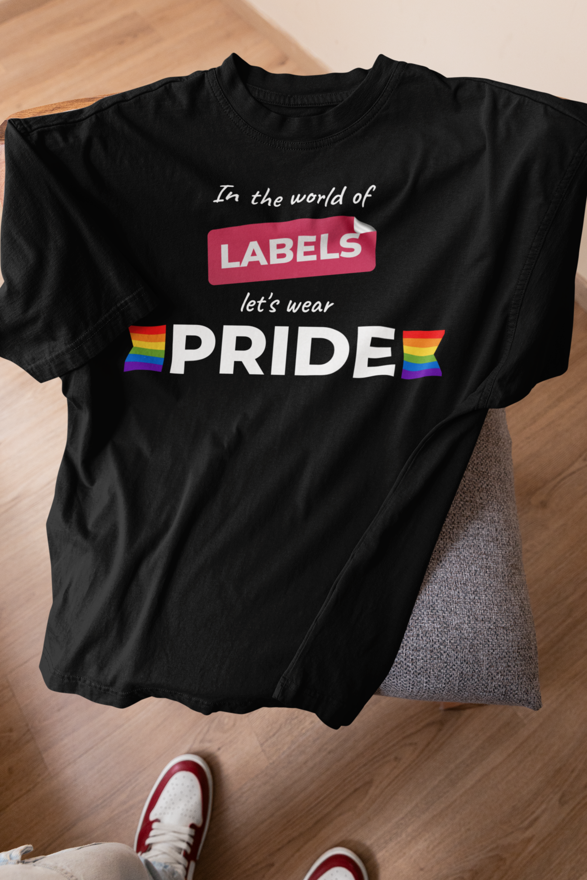 Black folded ANTI t-shirt featuring the slogan “In the world of labels, let’s wear PRIDE” in bold white text with rainbow flags, resting on a modern chair with sneakers nearby.

