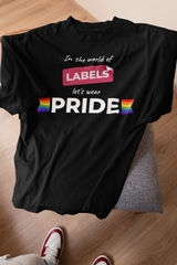 Black folded ANTI t-shirt featuring the slogan “In the world of labels, let’s wear PRIDE” in bold white text with rainbow flags, resting on a modern chair with sneakers nearby.

