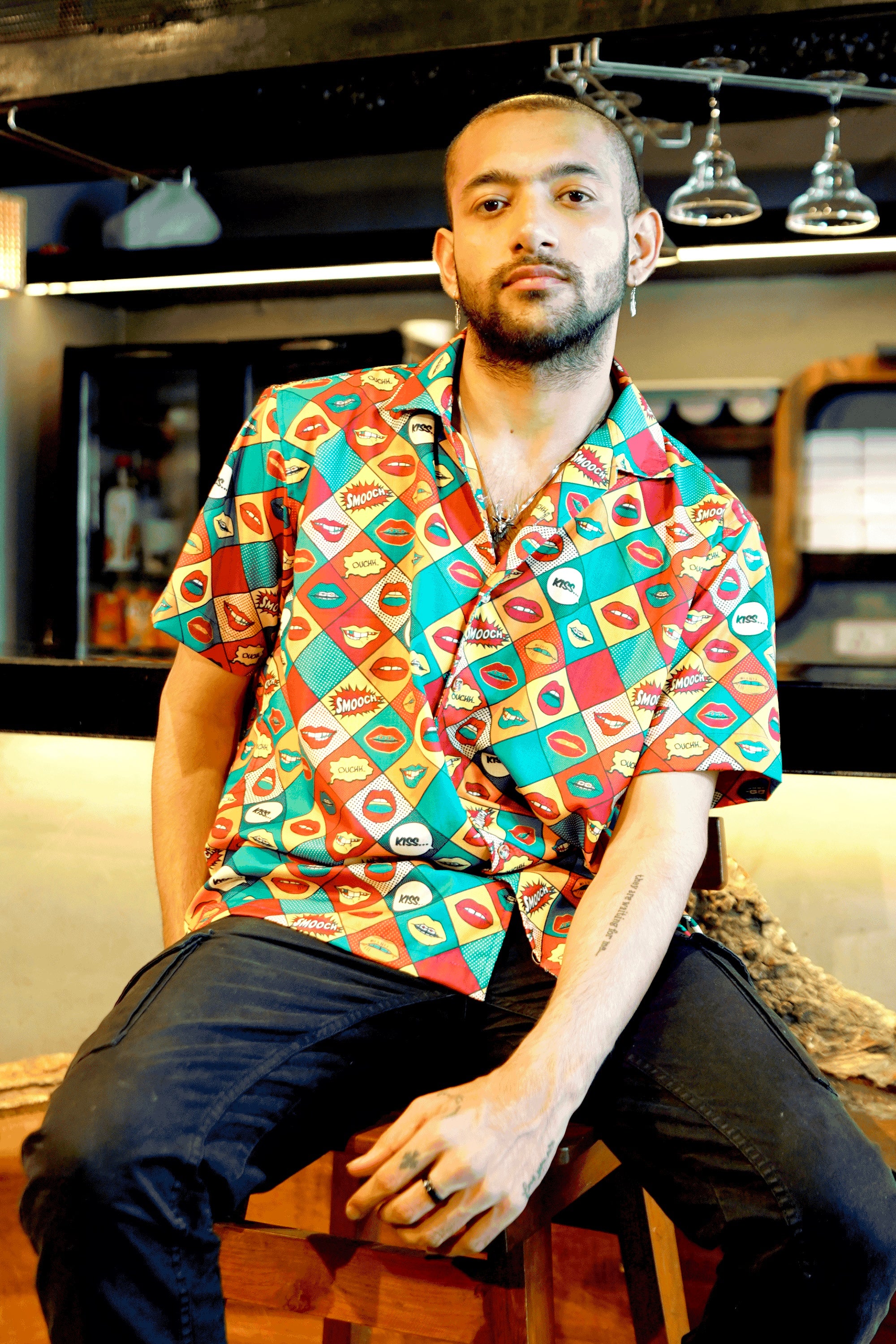 Detail of ANTI's vibrant short-sleeved shirt, highlighting its intricate pop art comic book print. The pattern features a kaleidoscope of colorful lips and expressive speech bubbles like 'KISS...' and 'SMOOCH,' set against a geometric background. The design is bold and playful, reflecting inclusive fashion.