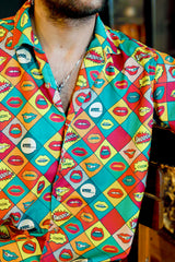A stylish gender-fluid person with a beard and light purple-tinted sunglasses sits confidently, wearing ANTI's short-sleeved button-up shirt featuring a vibrant pop art comic book style pattern of colorful lips and speech bubbles like 'KISS...' and 'SMOOCH.' This bold, expressive shirt embodies inclusive and playful fashion.
