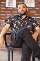 Full shot of a gender-fluid person sitting relaxed on a black chair against a rustic brick background, modeling ANTI's black short-sleeved shirt with a playful yet powerful 'love is love' and rainbow arc pattern. The look is casually bold and authentically expressive, celebrating LGBTQIA+ inclusivity.