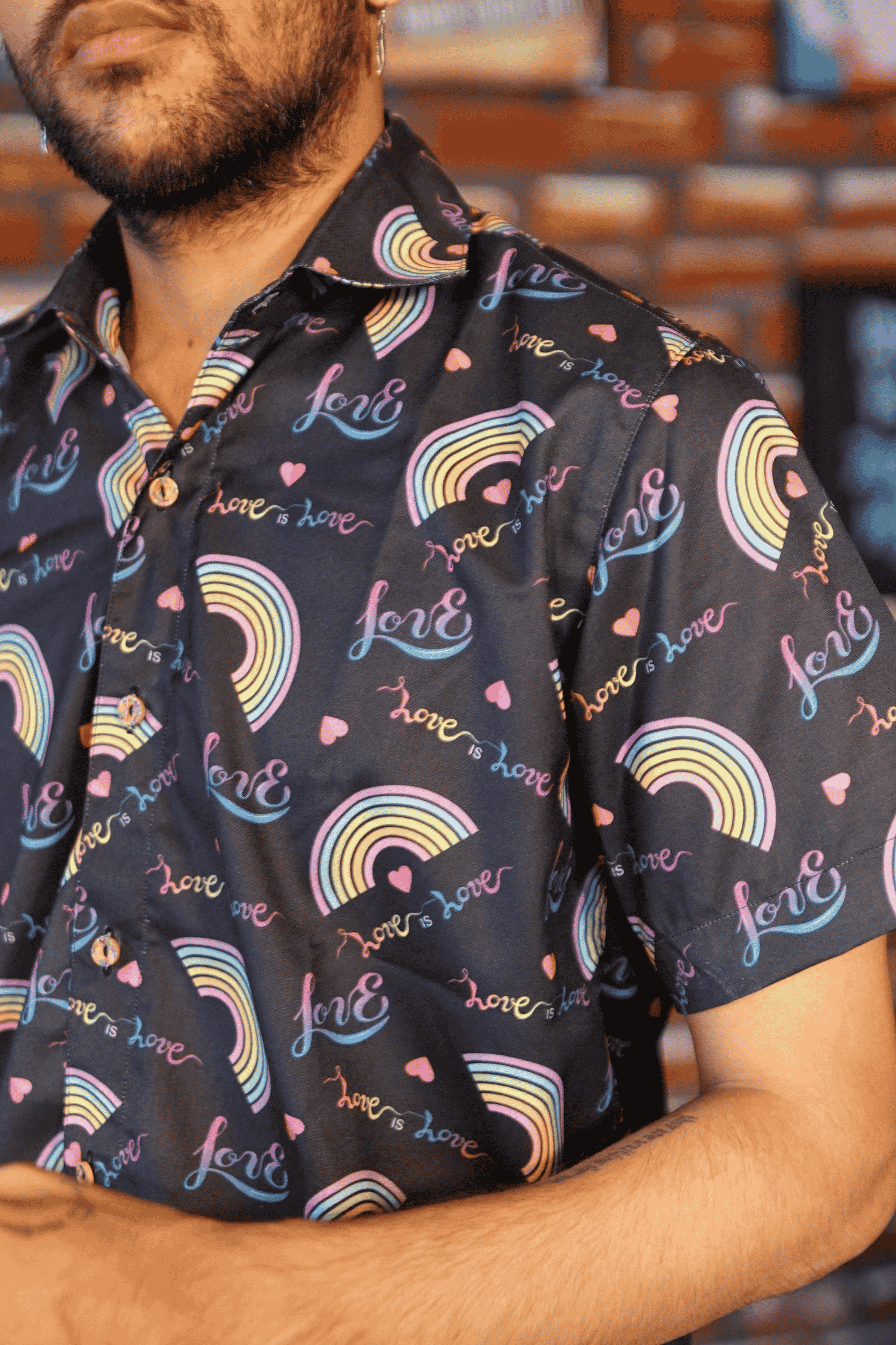 Close-up of a black short-sleeved shirt from ANTI, showcasing its detailed print of pastel rainbow arcs and handwritten-style 'love is love' text. The fabric appears soft and flowing, highlighting the inclusive and expressive design for LGBTQIA+ affirming fashion.