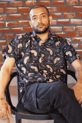 A gender-fluid person with a beard and tattoos sits confidently on a black chair against a brick wall, wearing a black short-sleeved shirt from ANTI, adorned with vibrant rainbow arcs and the text 'love is love.' The shirt embodies LGBTQIA+ pride and self-expression, perfect for inclusive fashion.