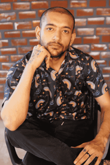 A gender-fluid individual with a shaved head, beard, and hand tattoos poses thoughtfully, hand on chin, while seated on a black chair. They are wearing ANTI's black short-sleeved shirt featuring a pattern of colorful rainbow arcs and 'love is love' text, symbolizing unity and queer pride.