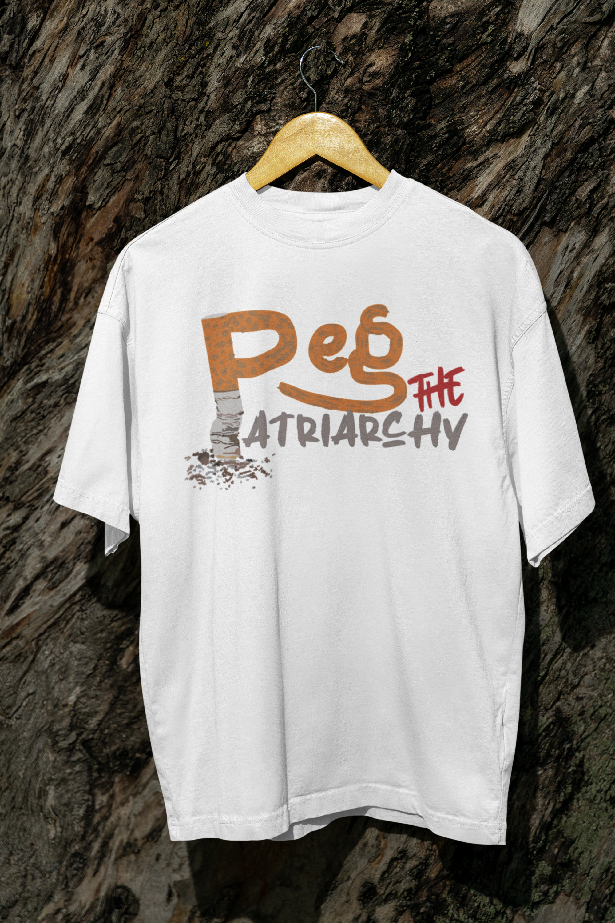 White gender-inclusive t-shirt hanging in front of a rugged wood backdrop, with a bold graffiti-style design reading “Peg the Patriarchy” in orange, red, and grey; the letter 'P' features a worn texture resembling a cigarette butt, symbolizing subversive anti-patriarchal sentiment.

