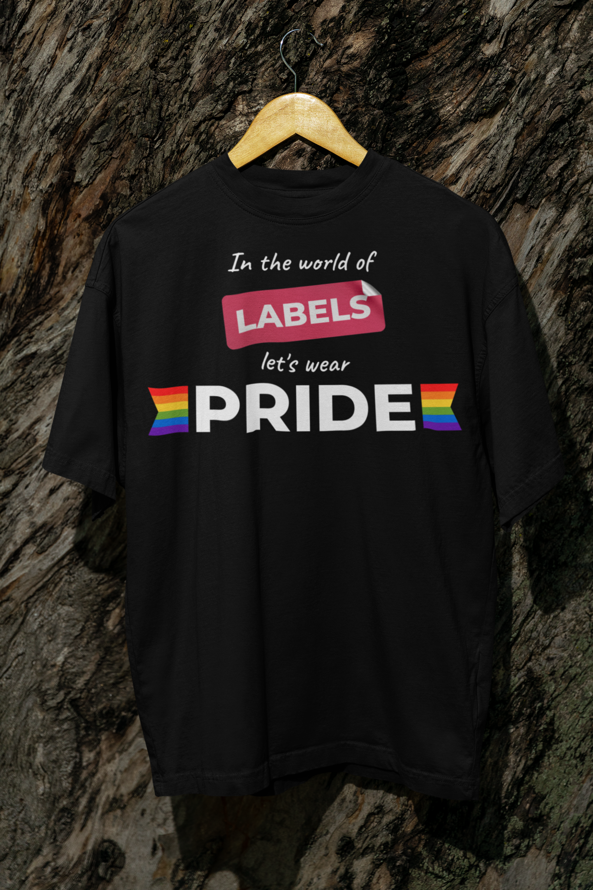 Black ANTI t-shirt with the slogan “In the world of labels, let’s wear PRIDE” and rainbow flag symbols, hanging on a wooden hanger against a tree trunk background.

