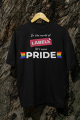 Black ANTI t-shirt with the slogan “In the world of labels, let’s wear PRIDE” and rainbow flag symbols, hanging on a wooden hanger against a tree trunk background.

