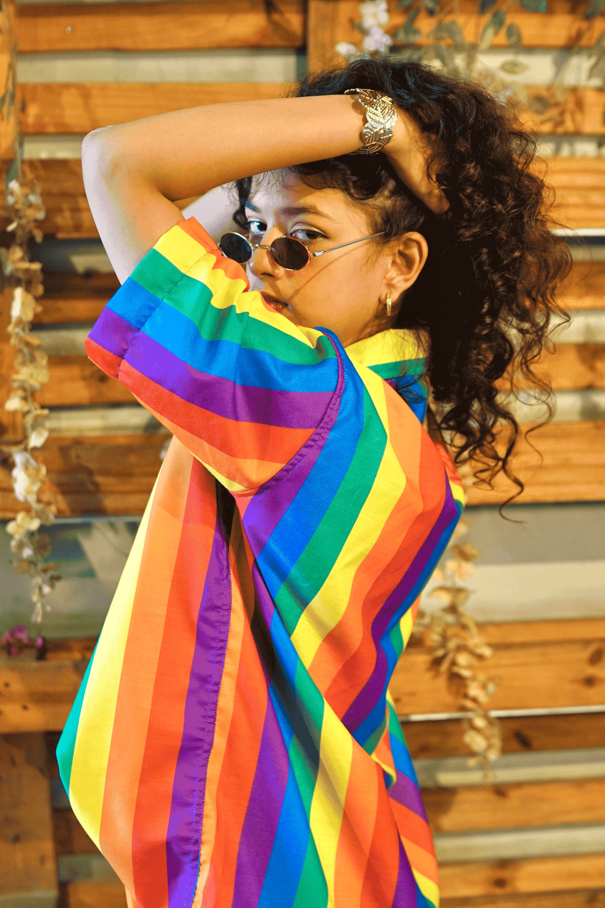 A gender-fluid person with curly hair and cool oval sunglasses confidently poses with their arm raised behind their head, looking back at the camera. They wear ANTI's vibrant short-sleeved button-up shirt featuring bold vertical rainbow stripes, symbolizing pride and inclusive style.