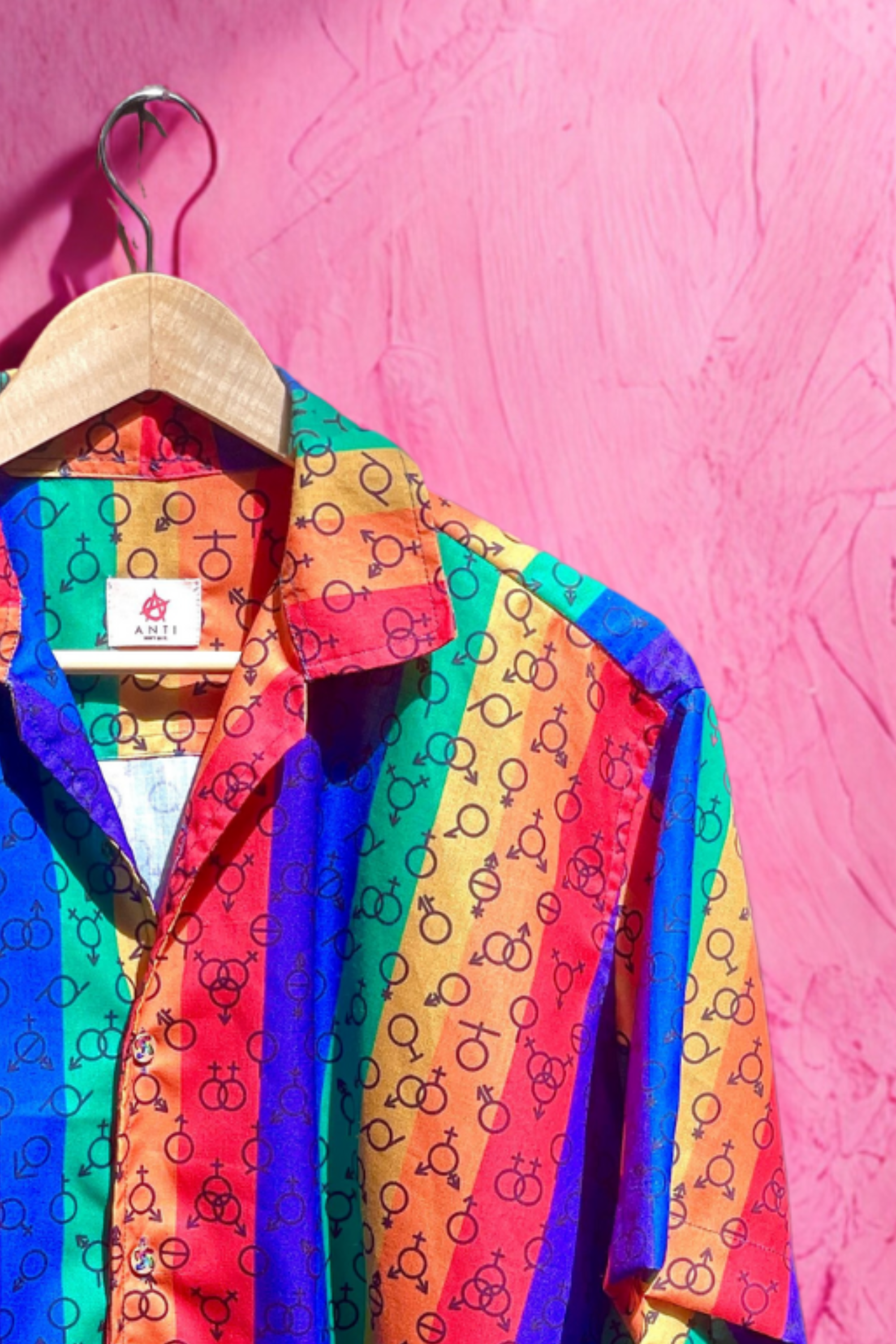 Colorful unisex short-sleeve button-up shirt featuring vertical rainbow stripes and an all-over pattern of LGBTQIA+ gender symbols, photographed against a textured pink wall. Designed by ANTI to celebrate gender diversity and queer identity.