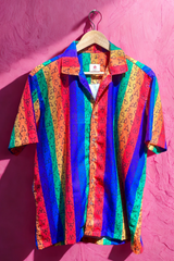 Colorful unisex short-sleeve button-up shirt featuring vertical rainbow stripes and an all-over pattern of LGBTQIA+ gender symbols, photographed against a textured pink wall. Designed by ANTI to celebrate gender diversity and queer identity.