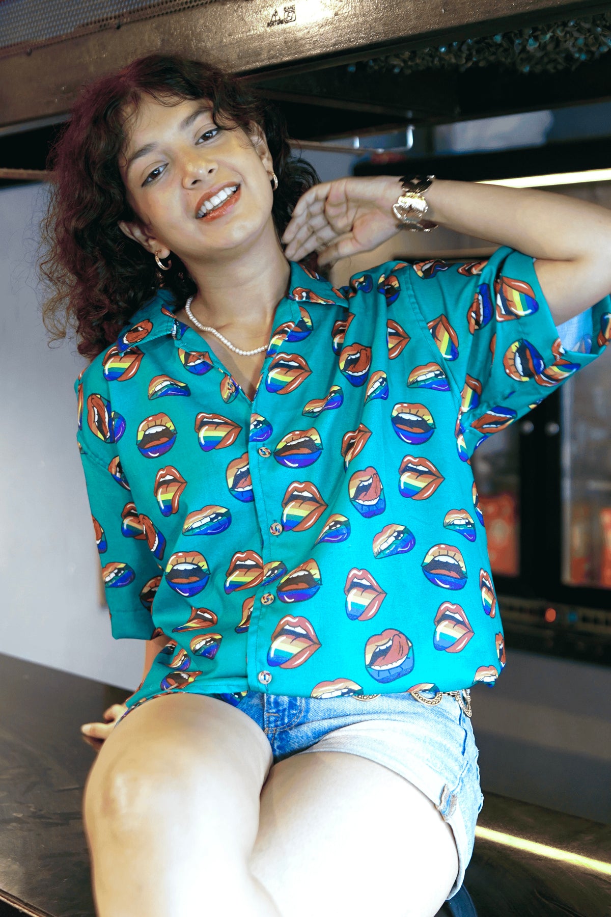 A dynamic shot of a gender-fluid person in a teal short-sleeved shirt with a vibrant lip pattern from ANTI, kneeling confidently on a surface with a neon 'drank' sign in the background. Paired with light wash denim shorts and a stylish belt, this look highlights the shirt's playful and inclusive design.