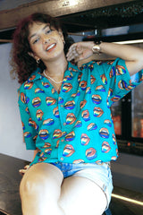 A dynamic shot of a gender-fluid person in a teal short-sleeved shirt with a vibrant lip pattern from ANTI, kneeling confidently on a surface with a neon 'drank' sign in the background. Paired with light wash denim shorts and a stylish belt, this look highlights the shirt's playful and inclusive design.