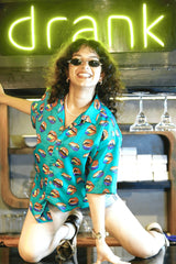 A joyful, gender-fluid person with curly hair and a pearl necklace smiles broadly, seated comfortably while adjusting their hair. They are wearing ANTI's distinctive teal shirt, which is adorned with an all-over pattern of colorful, open lips, capturing a playful and authentic spirit for inclusive fashion.