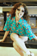 A confident, gender-fluid individual with curly hair and small oval sunglasses stands with hands in pockets, showcasing ANTI's eye-catching teal short-sleeved shirt. The shirt is covered in a repeated pattern of stylized, colorful open lips, representing a bold and expressive choice for any wardrobe.