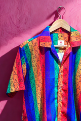 Short-sleeve button-up shirt with bold flame-inspired rainbow gradients and black gender symbols throughout, symbolizing fiery queer pride. Fashion-forward gender-fluid design displayed on a pink background for the brand ANTI.