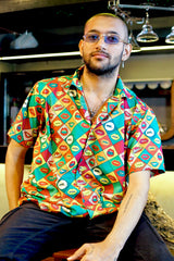 A confident gender-fluid model with a beard and minimal jewelry poses seated, showcasing ANTI's eye-catching short-sleeved shirt. The shirt displays a dynamic pop art grid pattern of various colored lips and playful text bubbles like 'SMOOCH' and 'OUCH!,' making it a unique statement piece for inclusive style.