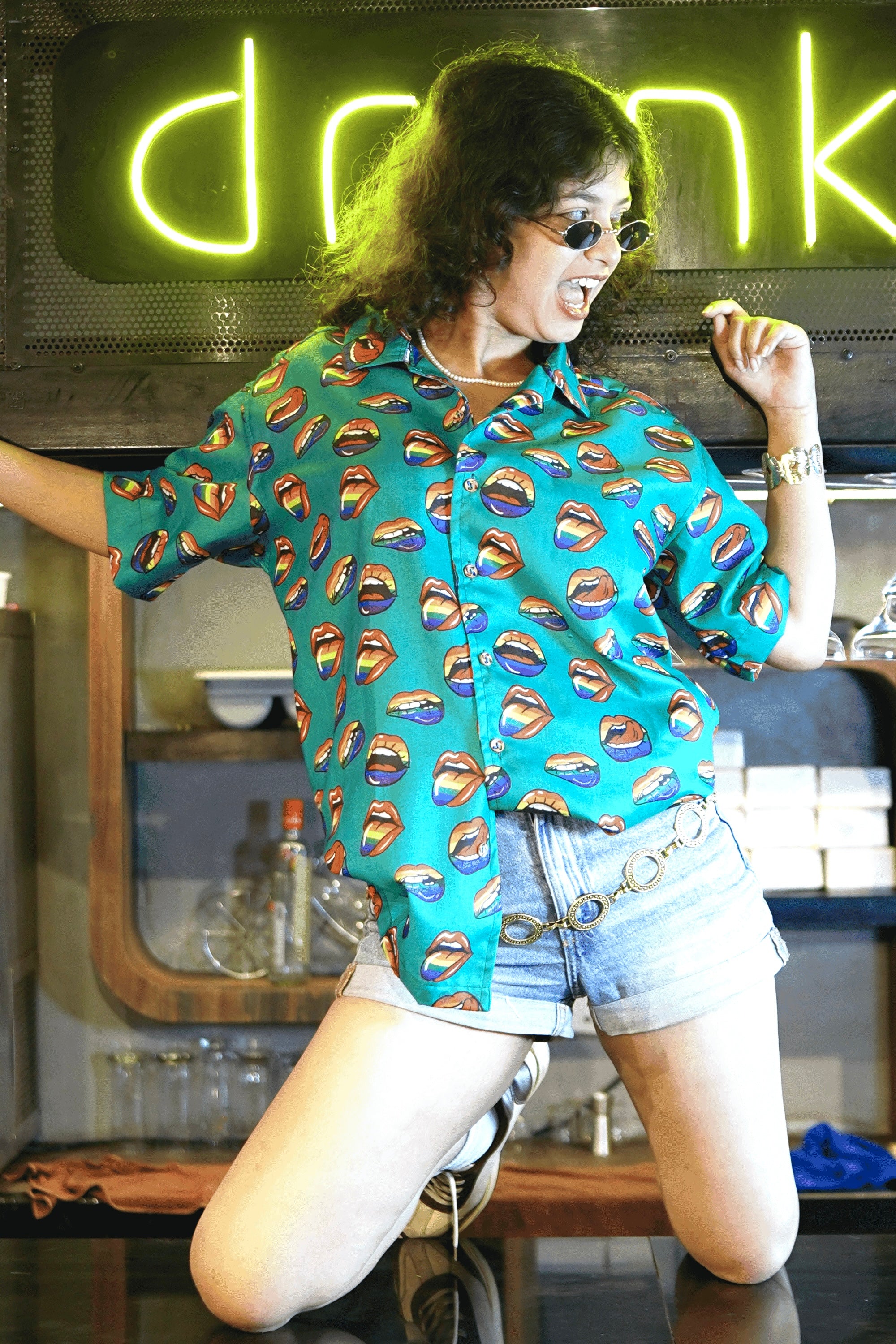 A playful, gender-fluid model with curly hair, wearing oval sunglasses, poses mischievously with their tongue out, seated on a dark counter. They're showcasing ANTI's unique teal shirt featuring a bold pattern of multi-colored open lips, exuding a confident and expressive vibe for inclusive style.
