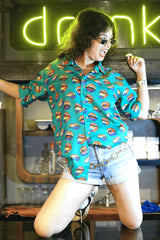 A playful, gender-fluid model with curly hair, wearing oval sunglasses, poses mischievously with their tongue out, seated on a dark counter. They're showcasing ANTI's unique teal shirt featuring a bold pattern of multi-colored open lips, exuding a confident and expressive vibe for inclusive style.