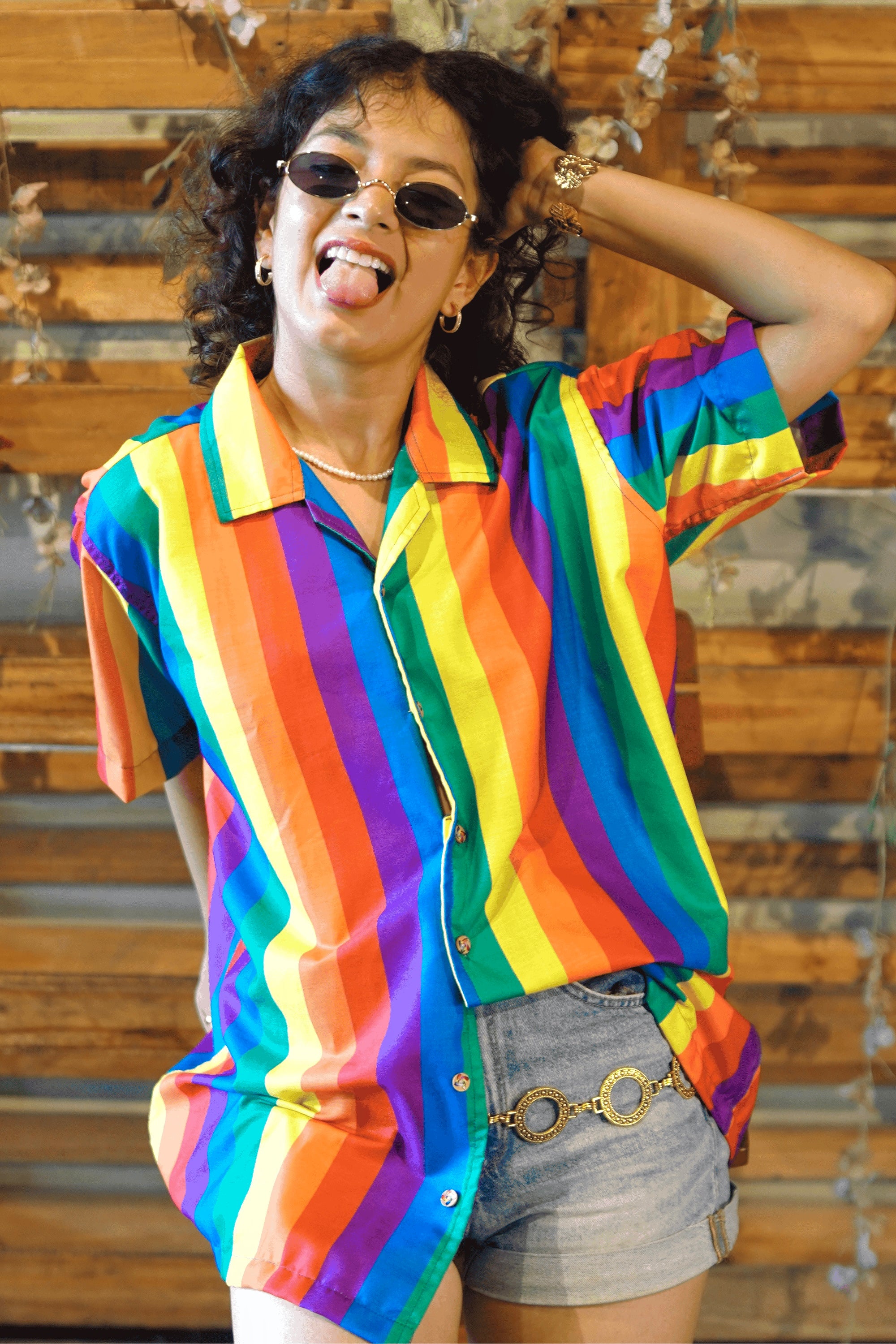 A gender-fluid model with curly hair and stylish sunglasses poses expressively with hands behind their neck, showcasing ANTI's striking short-sleeved shirt. The shirt boasts vibrant vertical rainbow stripes, paired with light-wash denim shorts and a gold chain belt, embodying a confident and inclusive fashion statement.