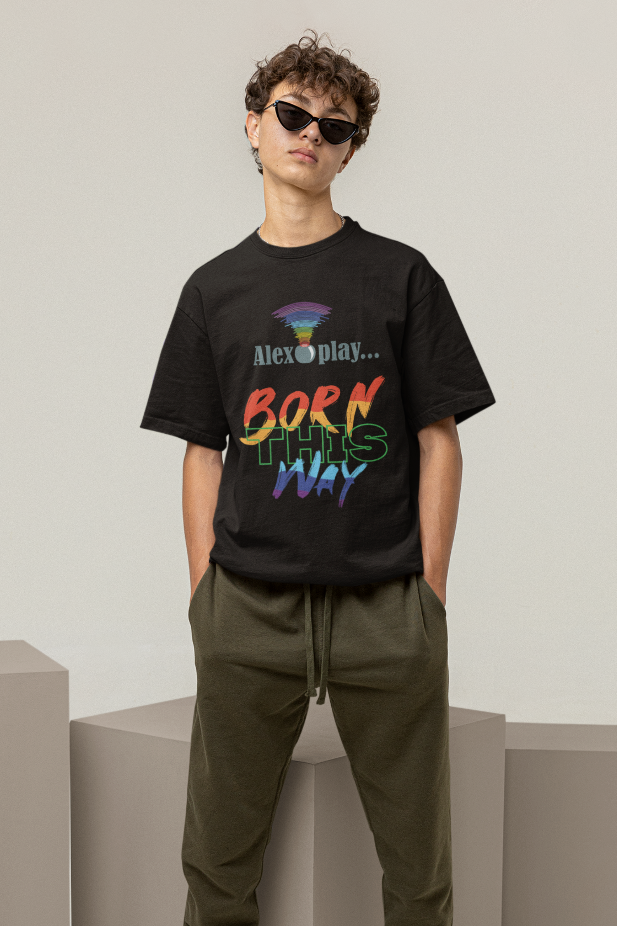 Model stands confidently in front of a neutral backdrop wearing the "Alexa, Play Born This Way" graphic T-shirt, styled in black with bold white typography across the chest. The shirt has a relaxed, oversized fit, symbolizing freedom of expression and body positivity. The model’s stance, with hands in pockets and gaze forward, exudes self-assurance and unapologetic queer pride. This look represents modern LGBTQ+ inclusive fashion with a streetwear edge.