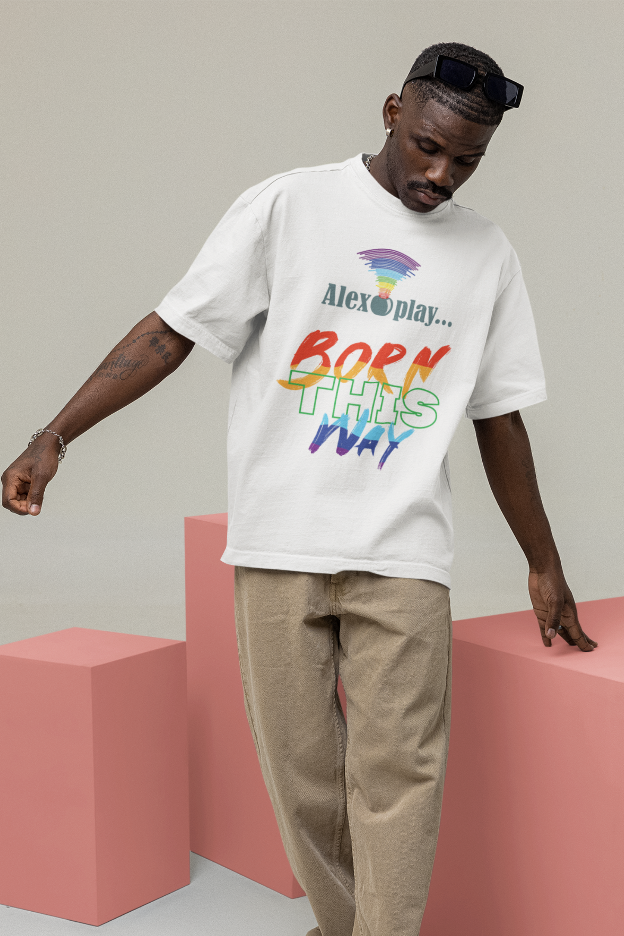 Close-up of the model seated in the "Alexa, Play Born This Way" T-shirt, showing off the sharp contrast of the white block text on black cotton fabric. The shirt’s androgynous silhouette and relaxed sleeves emphasize a gender-fluid fashion aesthetic. The model looks off-camera with a soft yet defiant expression, reinforcing the message of queer visibility and pride.