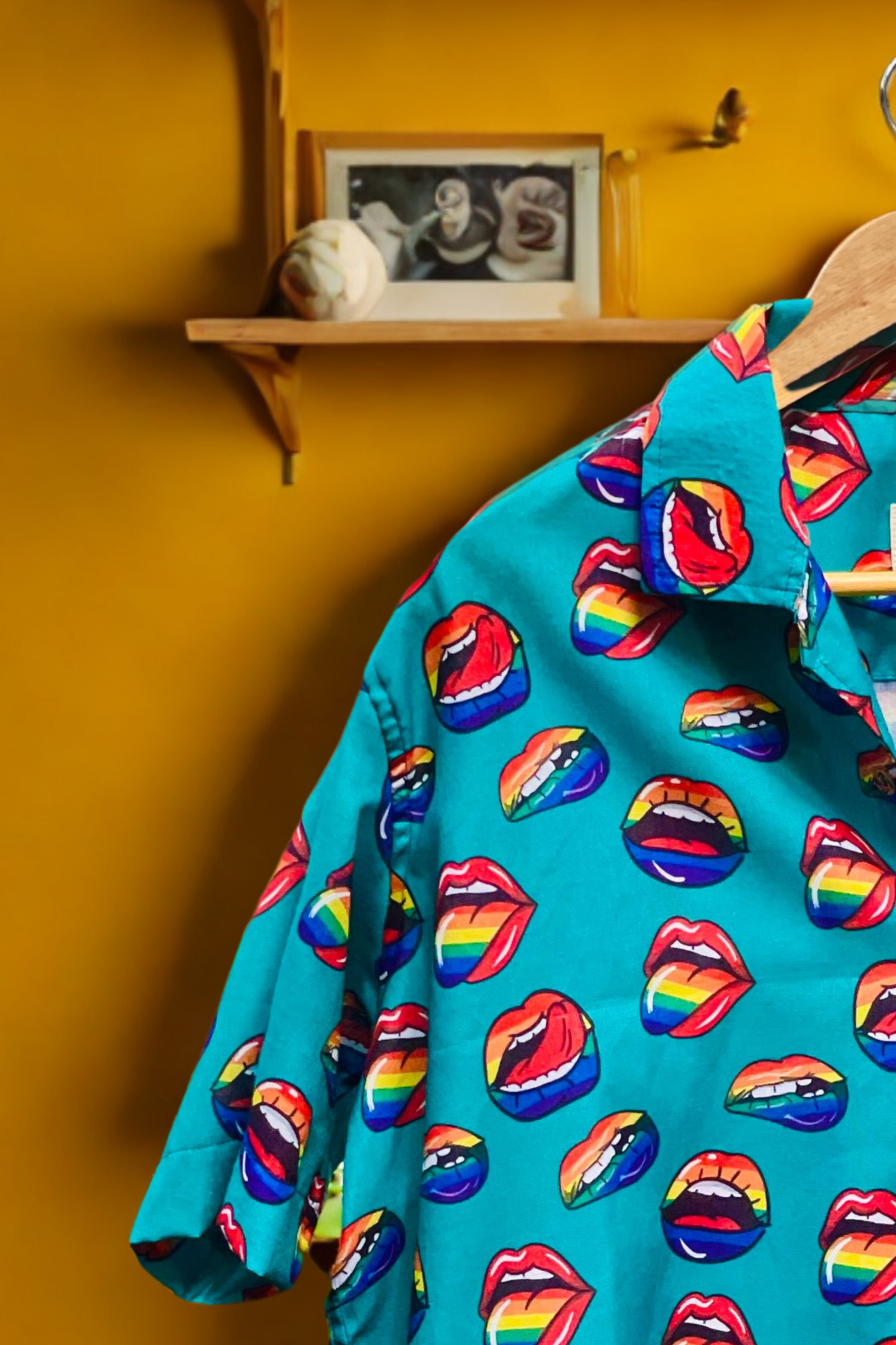 Flat-lay of a gender-neutral button-up shirt by ANTI, in a bold electric blue shade with an all-over print of biting lips in pink and red hues. The shirt channels sensual queer energy with a playful, rebellious edge. Featuring short sleeves, a structured collar, and breathable material, this shirt offers a street-savvy, unapologetically loud look for LGBTQIA+ individuals defying norms and embracing visibility.
