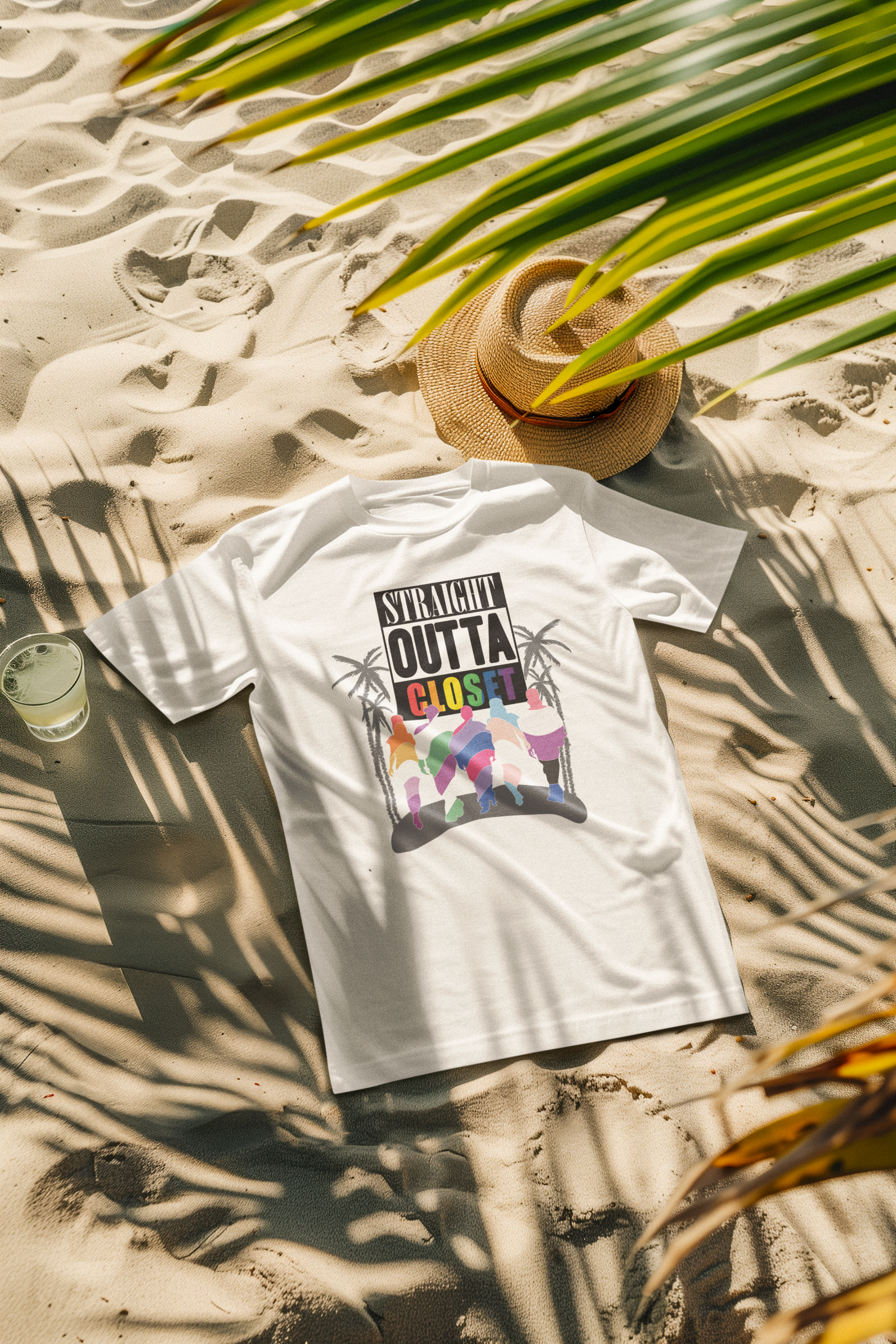 White "Straight Outta Closet" LGBTQ+ Pride T-shirt laid on sandy beach with a straw hat, palm leaves casting shadows, and a glass of lime water beside it.