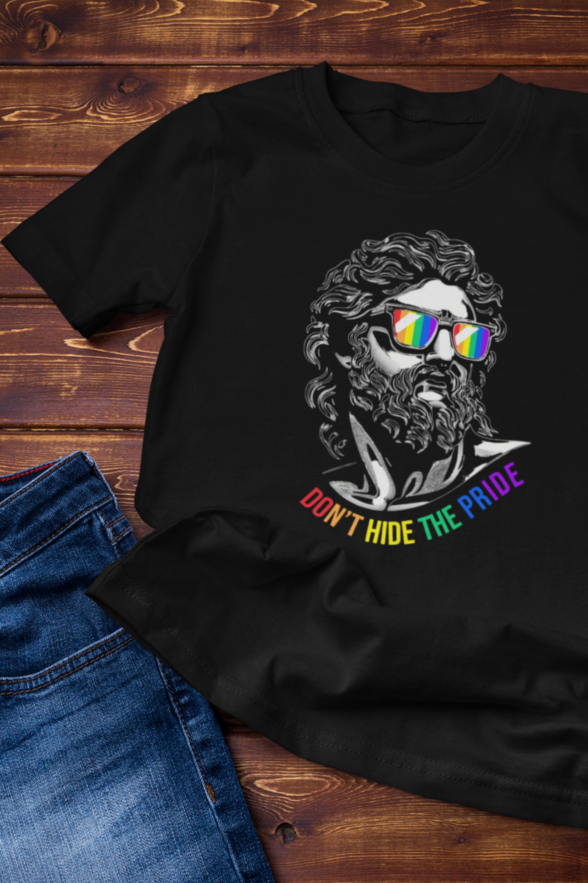 Flat lay of a black unisex t-shirt featuring a bold line-art sculpture graphic wearing rainbow-reflective sunglasses with the phrase "Don’t Hide The Pride" in rainbow text, styled with blue jeans on a wooden background — gender-fluid, queer-positive streetwear by ANTI.
