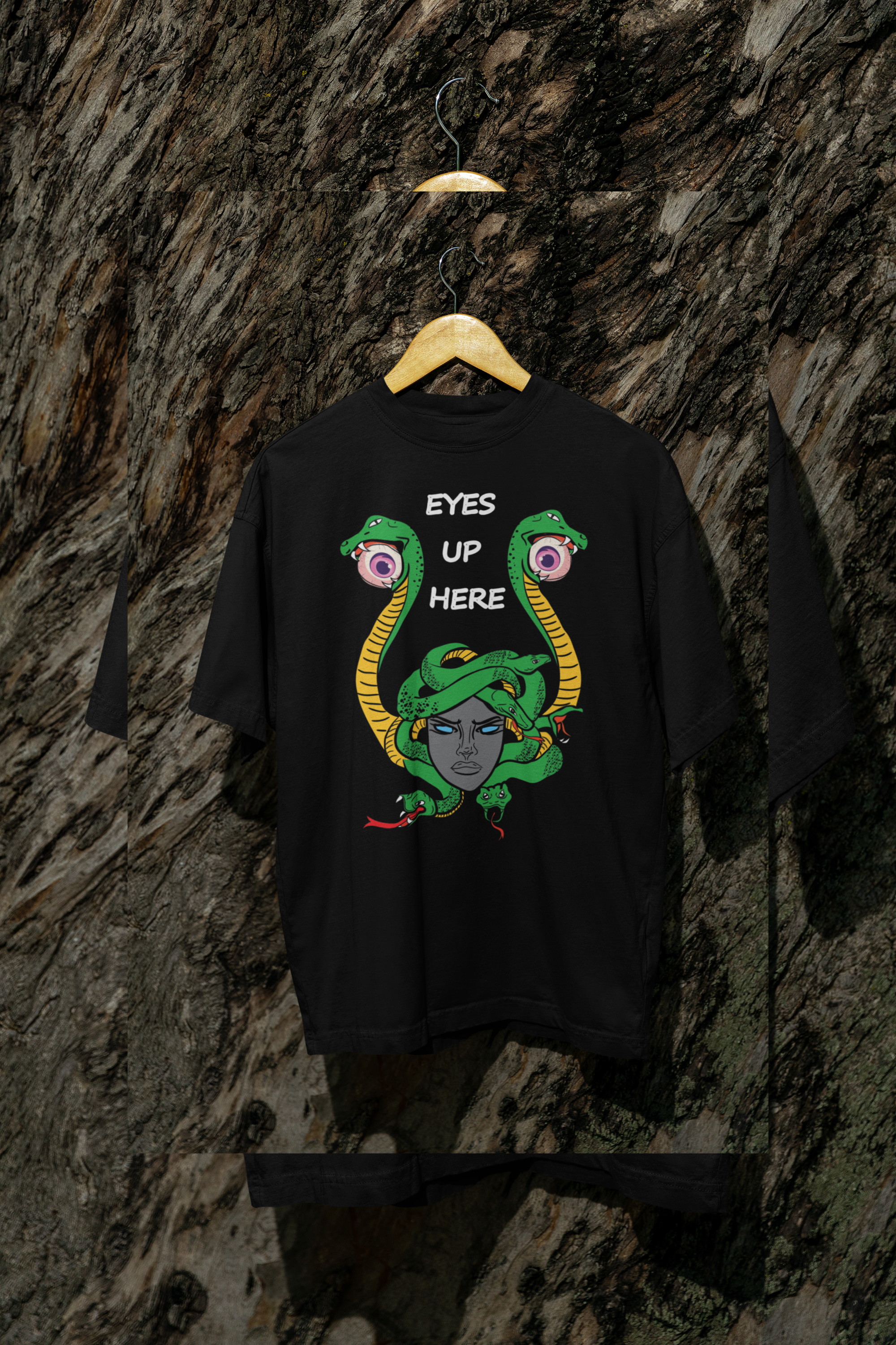 Black gender-fluid t-shirt featuring a bold Medusa-inspired graphic with green snakes, piercing blue eyes, and the phrase "EYES UP HERE" in white text, hanging on a wooden hanger against a textured tree bark background.