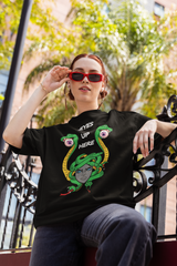 Bold gender-fluid model wearing a black oversized t-shirt with a vibrant Medusa illustration and the phrase “EYES UP HERE”; Medusa’s snakes feature eyeballs, styled with red sunglasses and streetwear attitude, framed by palm trees and urban architecture.