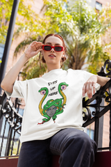 Confident, gender-fluid model wearing a white oversized t-shirt featuring a bold Medusa graphic with snakes holding eyeballs and the phrase “EYES UP HERE”; styled with red sunglasses and casual streetwear, set against a sunlit urban background with palm trees and wrought-iron details.