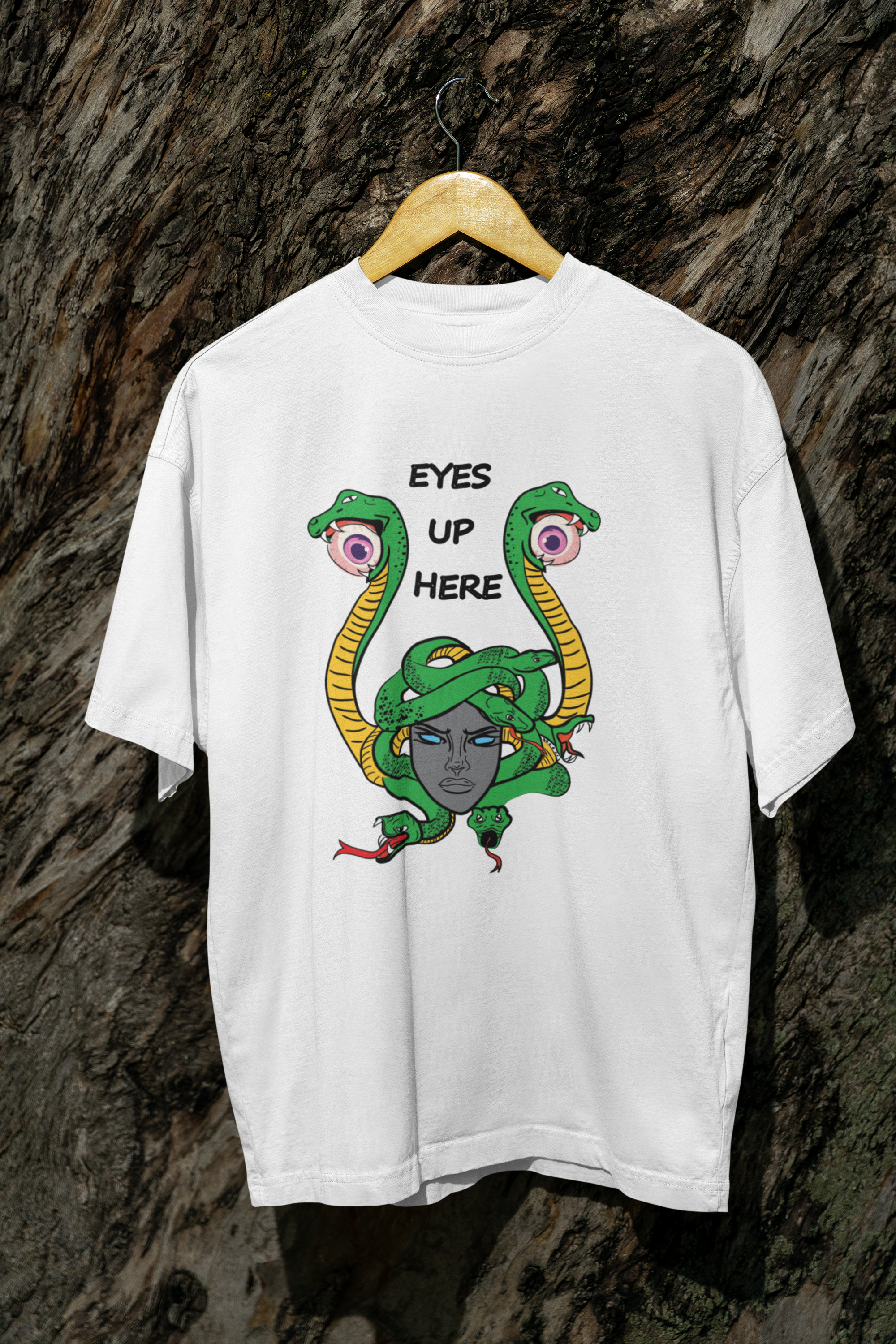 White streetwear tee with striking Medusa artwork in vibrant green, yellow, and gray tones and the phrase “EYES UP HERE,” displayed on a hanger against a textured bark wall, designed for gender-inclusive fashion expression.