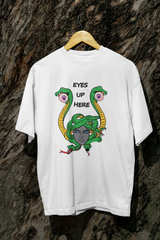 White streetwear tee with striking Medusa artwork in vibrant green, yellow, and gray tones and the phrase “EYES UP HERE,” displayed on a hanger against a textured bark wall, designed for gender-inclusive fashion expression.
