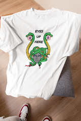 White unisex t-shirt featuring an expressive Medusa graphic with green serpents and floating eyes, plus bold “EYES UP HERE” text, laid on a gray chair over hardwood flooring with sneakers partially in frame.