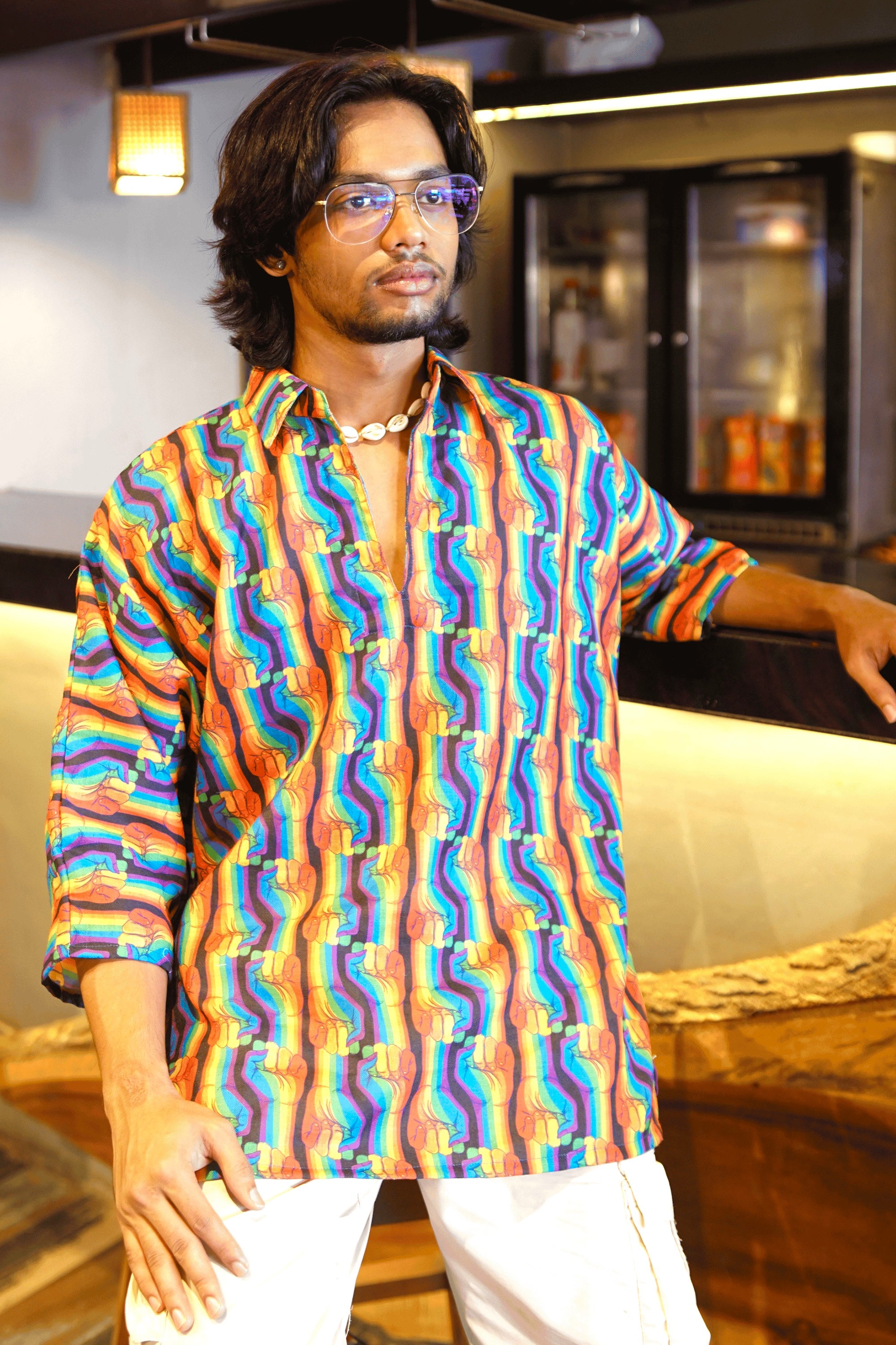 Person wearing a bold unisex shirt with rainbow protest fist print, styled with white cargos and shell necklace in indoor café setting.