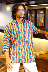 Person wearing a bold unisex shirt with rainbow protest fist print, styled with white cargos and shell necklace in indoor café setting.