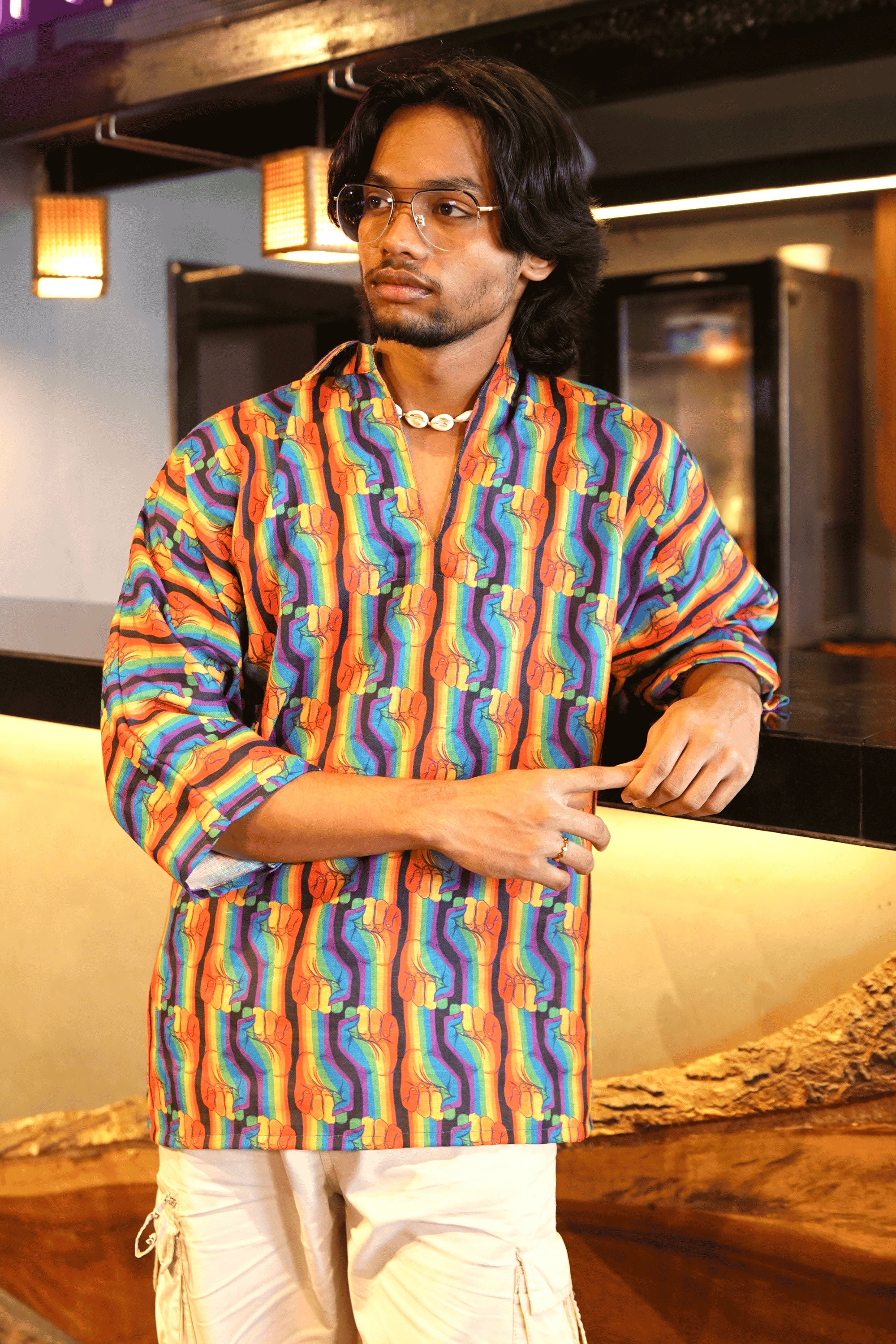 Close-up of person in rainbow fist print gender-neutral shirt with rolled-up sleeves, styled with white pants and minimal jewelry.