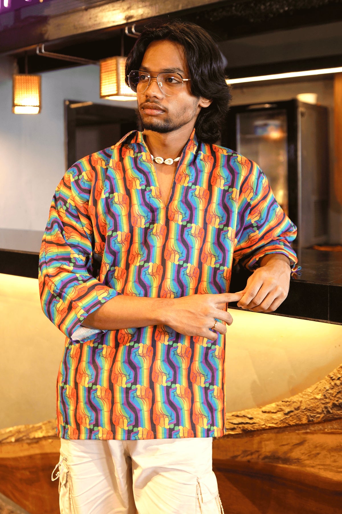 Close-up of person in rainbow fist print gender-neutral shirt with rolled-up sleeves, styled with white pants and minimal jewelry.