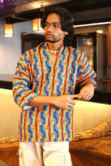 Close-up of person in rainbow fist print gender-neutral shirt with rolled-up sleeves, styled with white pants and minimal jewelry.