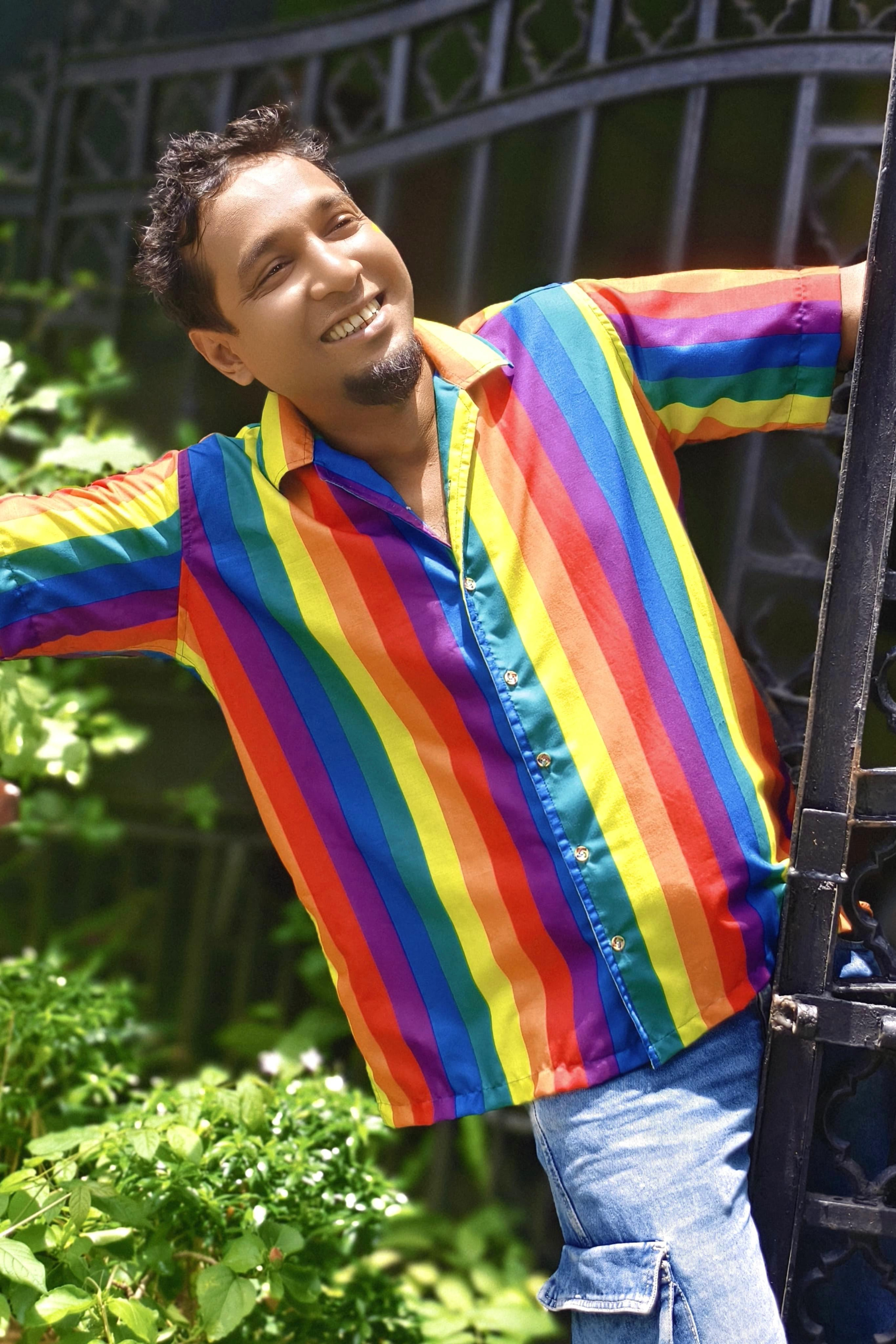 Individual outdoors wearing a rainbow-striped ANTI shirt, smiling with joy in a natural setting — celebrating queer freedom and soft rebellion.