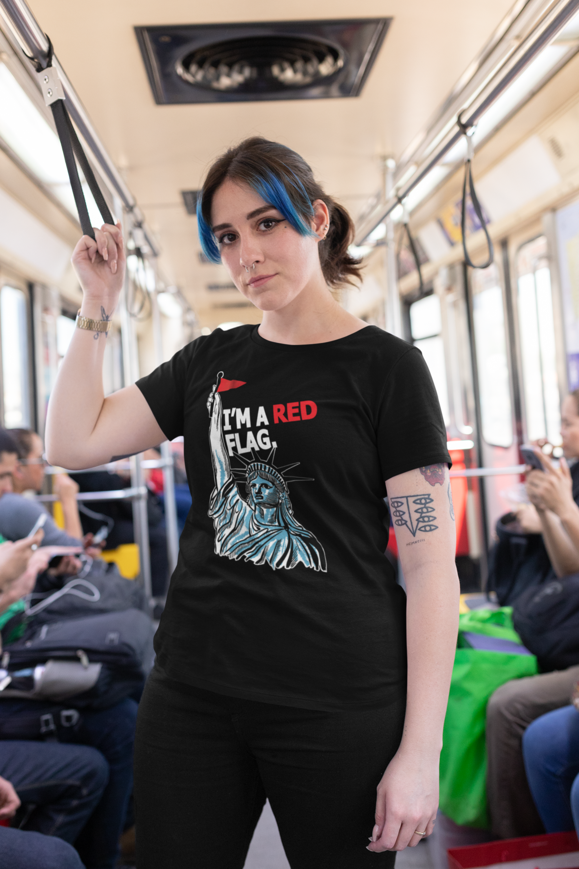  Person with blue highlights and a strong gaze wears a black inclusive graphic t-shirt with the Statue of Liberty raising a red flag and the phrase “I’M A RED FLAG”; urban subway background highlights ANTI’s genderless, rebellious pride apparel.

