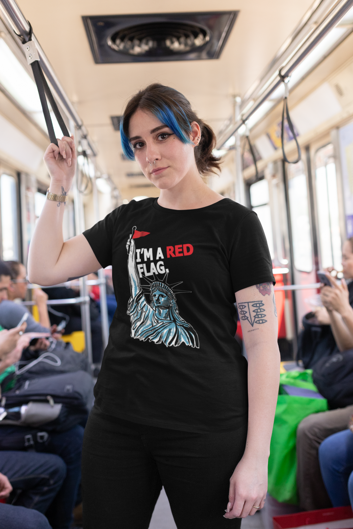 Person with blue highlights and a strong gaze wears a black inclusive graphic t-shirt with the Statue of Liberty raising a red flag and the phrase “I’M A RED FLAG”; urban subway background highlights ANTI’s genderless, rebellious pride apparel.

