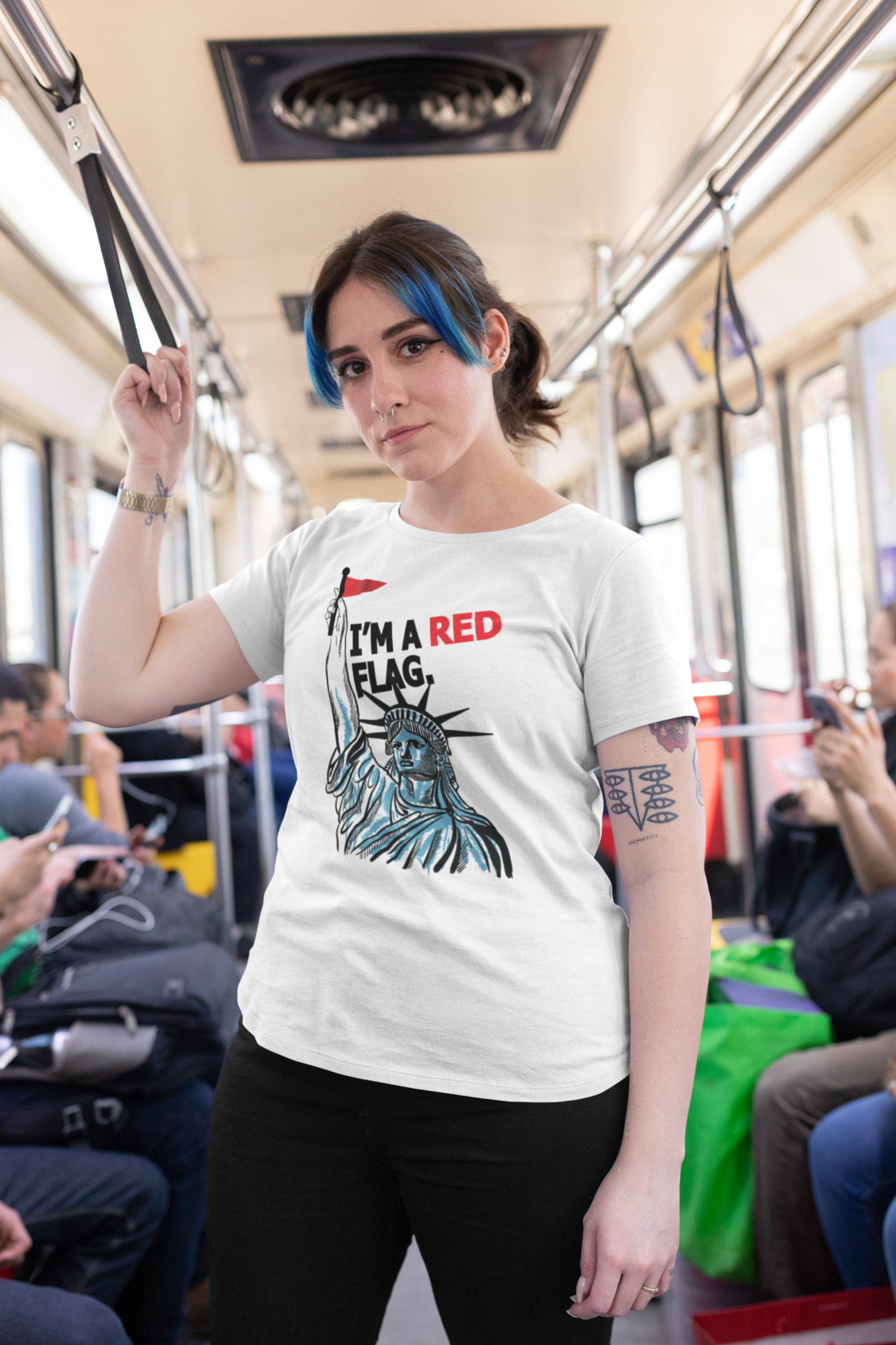 Confident person with blue hair and septum piercing wearing a white gender-fluid t-shirt featuring the Statue of Liberty holding a red flag with bold text saying “I’M A RED FLAG”; photographed on a public transit train, representing ANTI’s unapologetically queer streetwear fashion.
