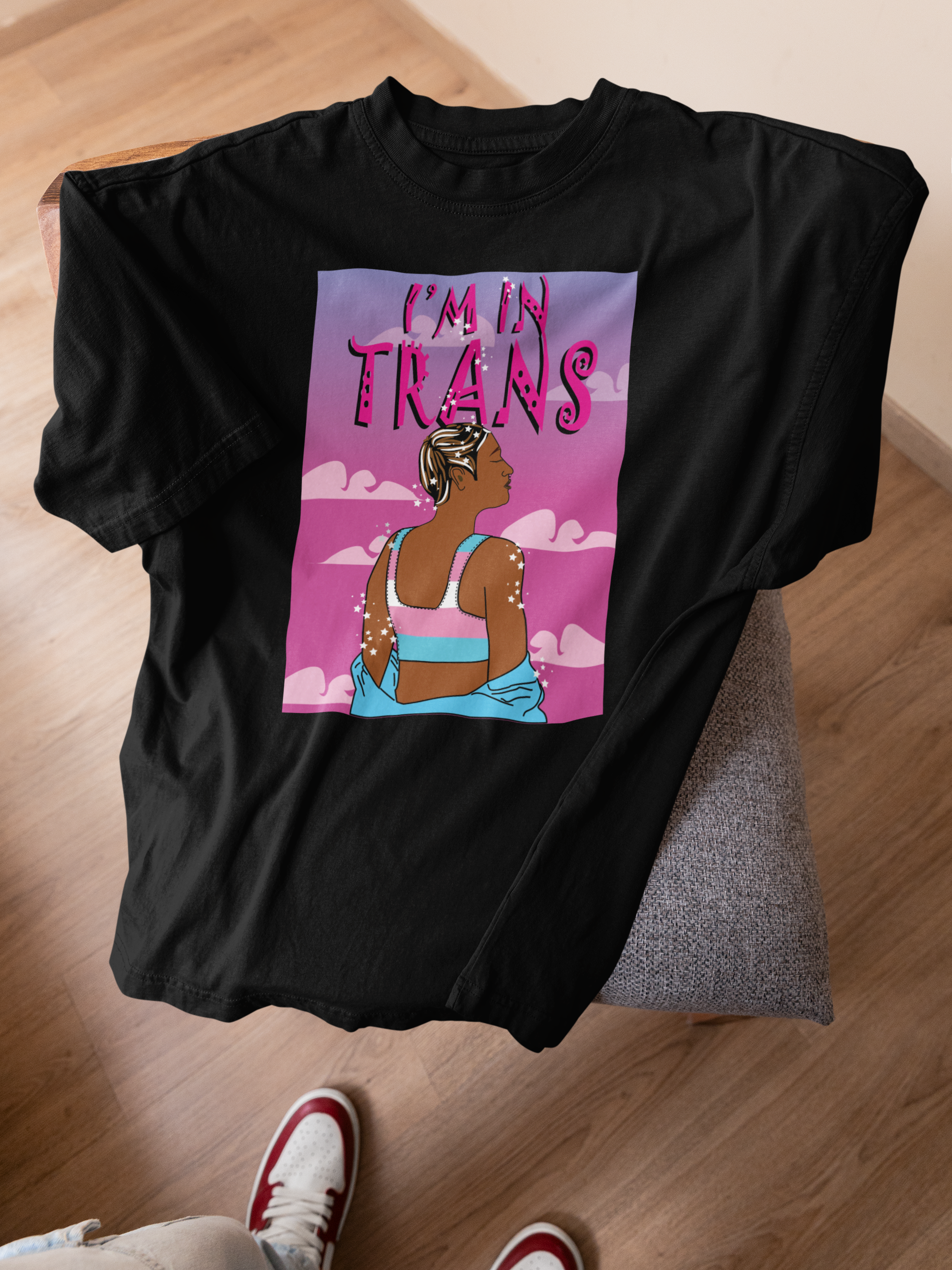 Black inclusive-fit t-shirt resting on a fabric chair, spotlighting a vibrant artwork of a trans individual from behind, confidently facing a pink sky with clouds, wearing a pastel binder in trans flag colors beneath the proud text “I’M IN TRANS.”